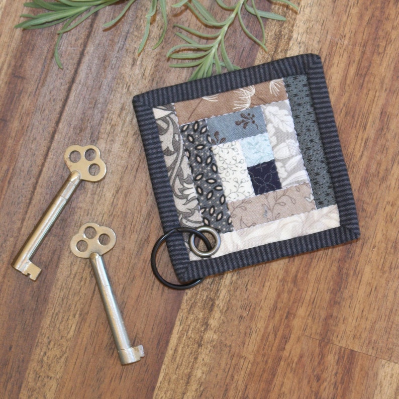Może przedstawiać: Mały, szary i biały patchworkowy koc z czarną obw&oacute;dką. Koc ma przymocowany brelok do kluczy. Dwa antyczne klucze leżą na drewnianej powierzchni.