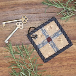 K&ouml;nnte beinhalten: Ein kleiner, quadratischer, gesteppter Schl&uuml;sselanh&auml;nger mit beigefarbenem Hintergrund und einem blauen und roten Kreuzmuster. Der Schl&uuml;sselanh&auml;nger hat einen schwarzen Rand und einen silbernen Metallring.