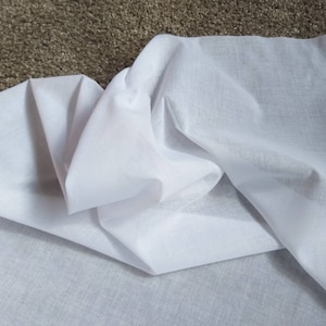 May include: White cotton fabric folded on a brown surface. The fabric is soft and smooth, with a slightly textured appearance.
