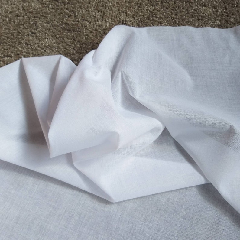 May include: White cotton fabric folded on a brown surface. The fabric is soft and smooth, with a slightly textured appearance.