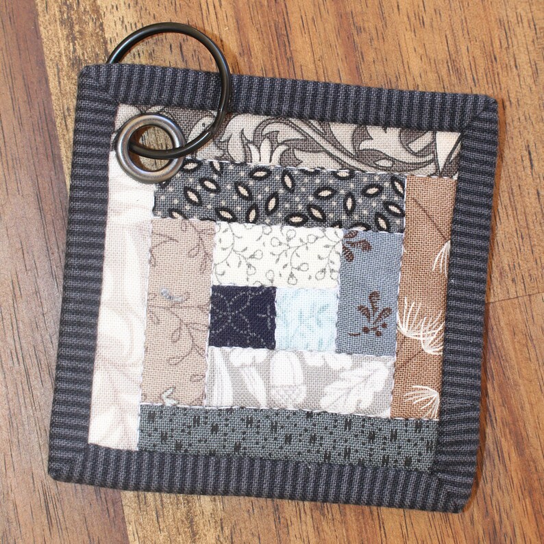 Może przedstawiać: Mały, szary, pikowany brelok do kluczy z czarnym metalowym k&oacute;łkiem. Pikowanie wykonane jest z r&oacute;żnych tkanin w odcieniach brązu, szarości i bieli.