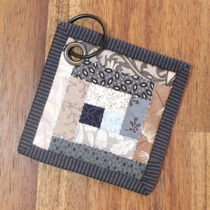 Może przedstawiać: Mały, kwadratowy, pikowany brelok do kluczy z wzorem z czarno-białej tkaniny. Brelok ma czarny metalowy pierścień przymocowany do g&oacute;ry.