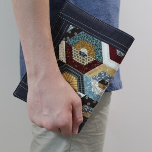 Puede incluir: Un embrague de tela azul oscuro con un diseño geométrico de patchwork con varios tonos de azul, marrón, amarillo y rojo. El embrague tiene un detalle de costura blanca alrededor de los bordes.