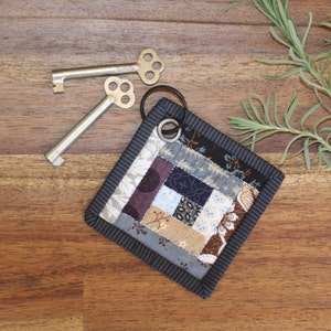 May include: A small, square, patchwork quilt keychain with a black and gray border. The quilt is made of various fabrics in shades of blue, brown, and purple. A silver key ring is attached to the keychain.