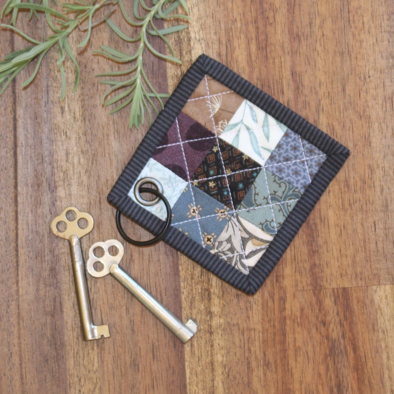 May include: A small, square, quilted fabric key holder with a black border and a patchwork design of various fabrics in shades of blue, brown, and purple. The key holder has a black metal ring attached to it.