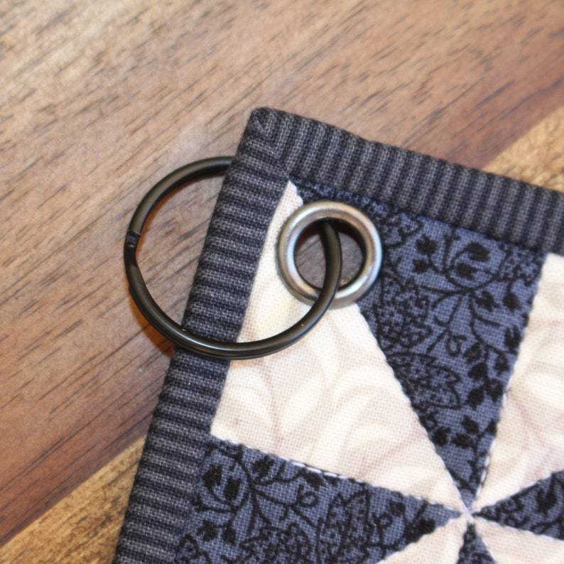May include: A black and white quilt with a grey fabric border and a black metal ring attached. The quilt has a geometric pattern with a floral design.