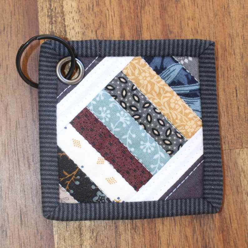 May include: A gray fabric key chain with a patchwork design featuring various shades of blue, brown, and yellow. The key chain has a metal ring attached.