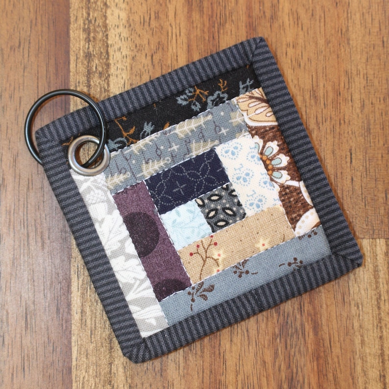 May include: A small, square, patchwork quilt keychain with a gray border and a metal ring. The quilt is made of various fabrics in shades of brown, blue, gray, and white.