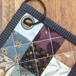 May include: A small, quilted key fob with a black metal ring. The fabric is a patchwork of different patterns in shades of blue, brown, and white.