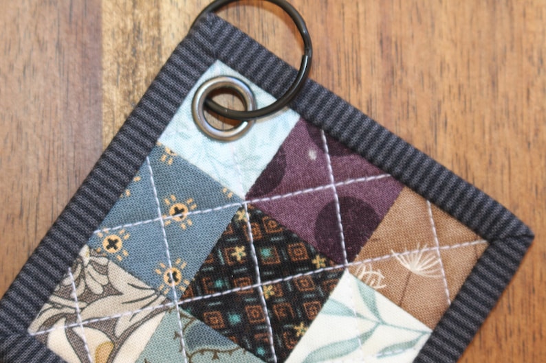 May include: A small, quilted key fob with a black metal ring. The fabric is a patchwork of different patterns in shades of blue, brown, and white.