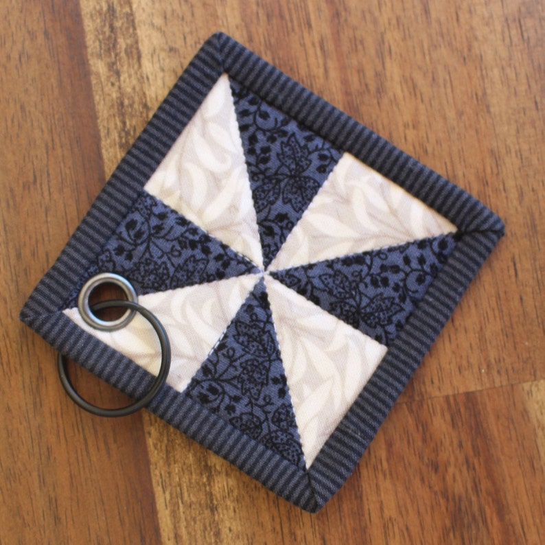 May include: A small, square, quilted fabric coaster with a blue and white geometric pattern. The coaster has a dark blue border and a metal ring attached to one corner.