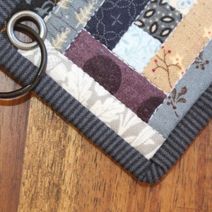 May include: A gray and black fabric key chain with a patchwork design and a metal ring. The fabric is made of different patterns and colors, including blue, white, and brown.