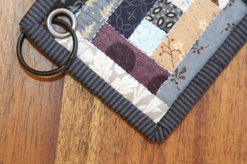 May include: A gray and black fabric key chain with a patchwork design and a metal ring. The fabric is made of different patterns and colors, including blue, white, and brown.