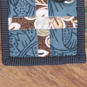 May include: A small, blue and brown fabric patchwork with a black border. The fabric has a floral pattern.