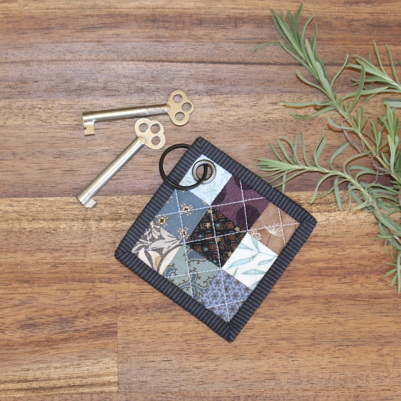 May include: A small, square, patchwork key fob with a black border and a silver key ring. The fob is made of different fabrics in shades of blue, brown, and white. The fabric is quilted and has a floral pattern.