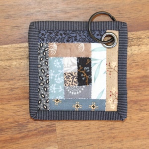 Peut inclure: Sous-verre carr&eacute; en tissu avec un motif patchwork dans les tons de bleu, marron, gris et cr&egrave;me. Le dessous de verre a une bordure ray&eacute;e bleu fonc&eacute; et un anneau m&eacute;tallique pour suspendre.