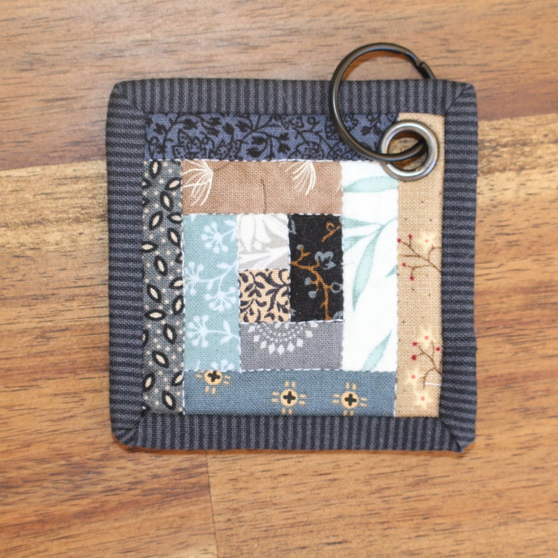 Peut inclure: Sous-verre carr&eacute; en tissu avec un motif patchwork dans les tons de bleu, marron, gris et cr&egrave;me. Le dessous de verre a une bordure ray&eacute;e bleu fonc&eacute; et un anneau m&eacute;tallique pour suspendre.