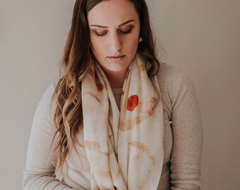 May include: A woman wearing a long-sleeved, light gray shirt and blue jeans. She is wearing a white scarf with brown and red abstract designs.