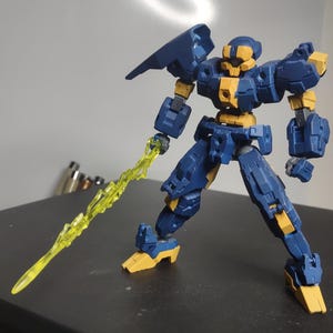 May include: A blue and yellow articulated robot model with a yellow energy sword. The robot has a detailed design with visible joints and panels. The model is posed on a black surface, showcasing its dynamic stance.