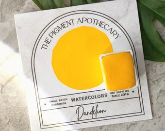 May include: A watercolor paint set featuring a vibrant yellow color labeled "Dandelion." The packaging includes the text "THE PIGMENT APOTHECARY," "WATERCOLORS," and "ART SUPPLIES SINCE 2018." The set is small batch and handmade.