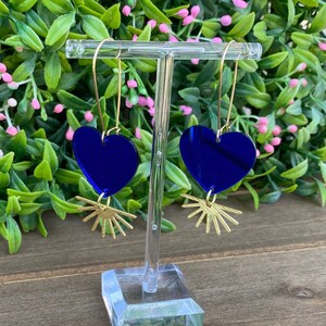 May include: A pair of blue heart-shaped earrings with gold sunburst accents. The earrings are hanging from a gold hook.
