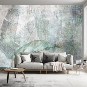 Könnte beinhalten: Ein hellgraues Sofa mit einer weißen Decke und zwei Kissen steht vor einer Wand mit einem abstrakten Muster in Grün und Grau. Das Muster zeigt ein geometrisches Design mit einem verblassten, Vintage-Look.