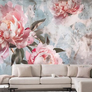 Op de afbeelding: Een vintage-stijl bloemenbehang met grote roze pioenen met groene bladeren op een lichtblauwe en grijze textuur achtergrond. Een witte hoekbank staat op de voorgrond.