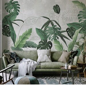Op de afbeelding: Een groene bank met kussens en een deken in een woonkamer met een behang met tropische bladmotieven. Een kleine tafel met een vaas en een plant erop staat voor de bank.
