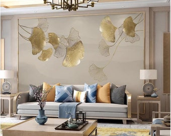 Op de afbeelding: Een woonkamer interieur met een lichtgrijze bank met blauwe en oranje kussens. De muur achter de bank heeft een grote gouden metalen wandkunst met een ontwerp van gestileerde ginkgo bladeren. De kamer is ingericht met een vaas met roze bloemen, een salontafel en een lamp op een bijzettafel.