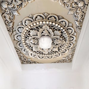 Op de afbeelding: Een wit plafond met een decoratief, versierd, goudkleurig, rond medaillon met een kristallen kroonluchter in het midden.