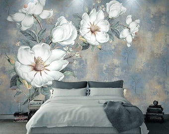 May include: A white floral wallpaper mural with a distressed blue background. The mural features large white magnolia flowers with green leaves. The mural is displayed in a bedroom setting with a bed and a nightstand.