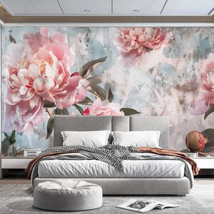 Op de afbeelding: Een slaapkamerinterieur met een grote bloemenmuurschildering met roze pioenen op een lichtblauwe en grijze achtergrond. De muurschildering bevindt zich achter een wit bed met grijs beddengoed en een grijze voetenbank.