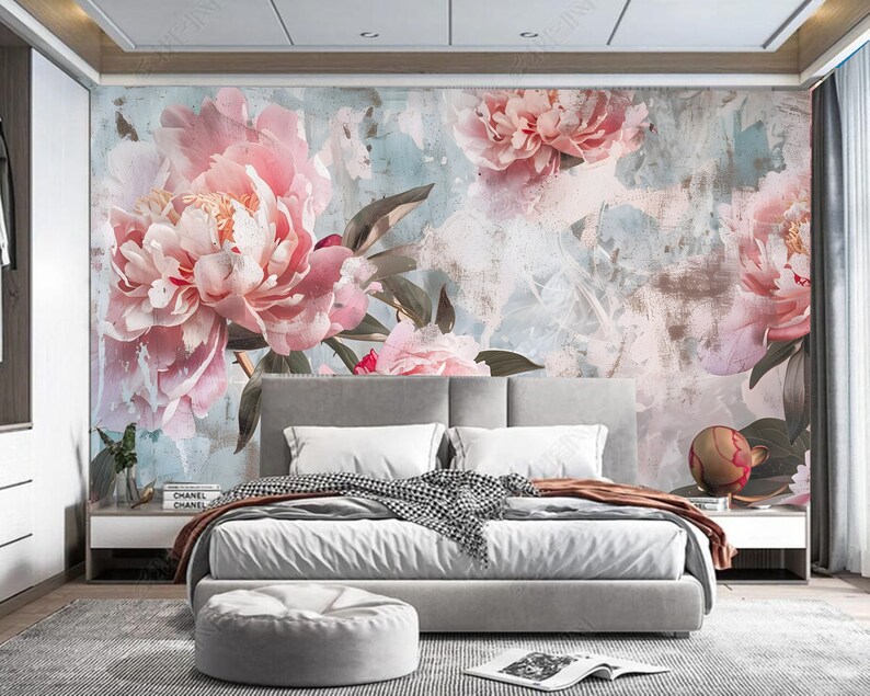 Op de afbeelding: Een slaapkamerinterieur met een grote bloemenmuurschildering met roze pioenen op een lichtblauwe en grijze achtergrond. De muurschildering bevindt zich achter een wit bed met grijs beddengoed en een grijze voetenbank.