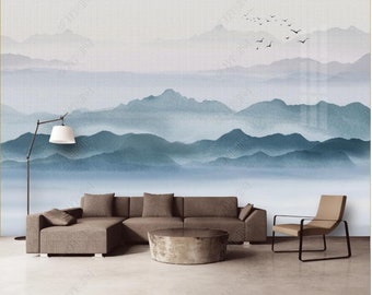 May include: A watercolor painting of a mountain range with a soft blue and gray color scheme. The mountains are layered and appear to be in the distance. There are small black birds flying in the sky above the mountains. The painting is displayed on a wall in a living room setting with a brown sectional sofa, a coffee table, and a chair.