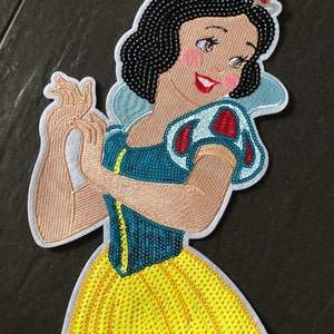 Könnte beinhalten: Ein gestickter Aufnäher von Schneewittchen aus Disneys Schneewittchen und die sieben Zwerge. Der Aufnäher zeigt Schneewittchen in ihrem ikonischen gelben Kleid mit einem blauen Oberteil und roten Ärmeln. Sie trägt eine Krone und lächelt strahlend.