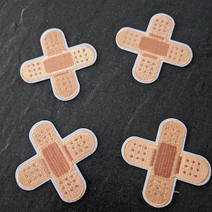 Könnte beinhalten: Vier gestickte Bügelbilder in Form von Pflastern in hellbrauner Farbe.