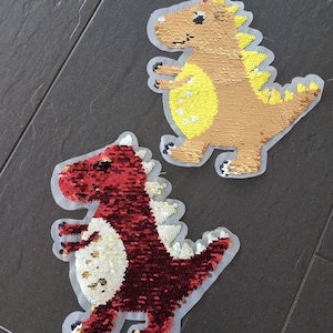 Könnte beinhalten: Zwei Pailletten-Dinosaurier-Aufnäher, einer rot und einer braun, mit gelben Akzenten. Die Aufnäher befinden sich auf einer grauen Fliesenfläche.