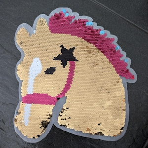 Könnte beinhalten: Eine goldene und rosafarbene Pailletten-Pferdekopf-Applikation mit einem schwarzen Stern auf der Stirn. Das Pferd hat ein rosafarbenes und weißes Zaumzeug.