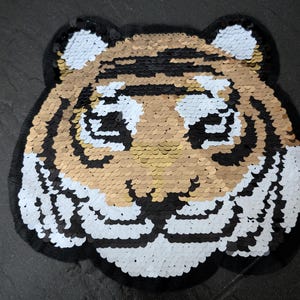 Könnte beinhalten: Ein goldfarbener, schwarzer und weißer Pailletten-Tigerkopf-Patch. Der Tiger hat eine schwarze Nase und schwarze Augen.