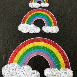 Könnte beinhalten: Drei regenbogenfarbene Pailletten-Patches mit weißen Wolkenformen. Die Regenbögen sind übereinander gelegt, wobei der kleinste Regenbogen oben und der größte unten liegt.