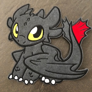 Könnte beinhalten: Eine schwarze und rote Filzapplikation von Ohnezahn, dem Nachtfurien-Drachen aus der Filmreihe Drachenzähmen leicht gemacht. Der Drache sitzt mit ausgebreiteten Flügeln. Er hat gelbe Augen und eine rote Schwanzflosse.