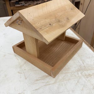 Puede incluir: Comedero para pájaros de madera con techo inclinado y base de bandeja. El comedero está construido con madera de color claro y presenta un anillo de metal para colgarlo. Diseño sencillo y funcional.