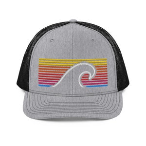 May include: A gray trucker hat with a black mesh back. The front features a colorful embroidered design of a wave over a striped sunset in shades of yellow, orange, red, pink, and blue. The hat has a curved brim.