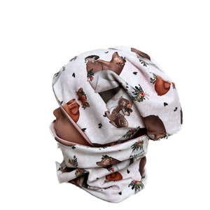 Könnte beinhalten: Eine rosa-weiße Strickmütze und ein passender Halsschmuck mit einem Waldtier-Druck. Der Druck zeigt Hirsche, Eichhörnchen und Bären, die Blumenkränze tragen.