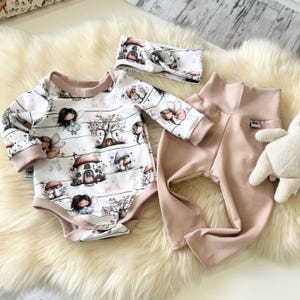 Pode incluir: Um conjunto de roupa de bebé com um body branco com estampa de fadas e cogumelos, uma faixa de cabeça a condizer e calças cor-de-rosa. O body tem mangas compridas e uma borda castanha clara. Um pequeno animal de pelúcia também é visível.