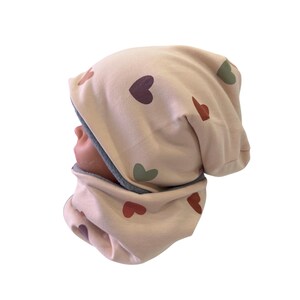 Może przedstawiać: Jasnoróżowy zestaw czapki i komina. Czapka i komin są ozdobione sercami w odcieniach bordowego, zielonego i czerwonego. Wnętrze czapki i komina jest szare.