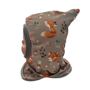 Könnte beinhalten: Graues Baby-Set aus Mütze und Halstuch mit einem sich wiederholenden Muster aus orangefarbenen Füchsen, Pilzen und Blättern. Die Mütze hat eine spitze Oberseite und eine graue Einfassung um das Gesicht und das Halstuch.