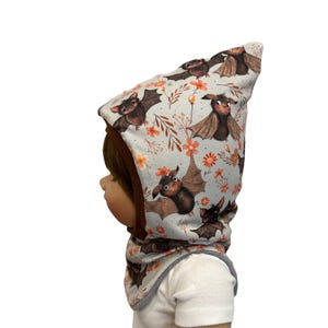 Könnte beinhalten: Hellblaues Baby-Set aus Mütze und Halstuch. Der Stoff hat ein sich wiederholendes Muster aus braunen Fledermäusen, orangefarbenen Blumen und grünen Blättern. Die Mütze hat eine braune Bordüre und wird von einer Puppe getragen.