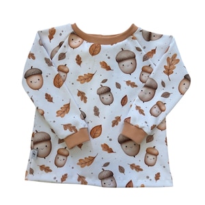 Könnte beinhalten: Weißes Langarmshirt mit einem Muster aus lächelnden Eicheln und braunen Blättern. Das Shirt hat braune Abschlüsse an Ausschnitt und Ärmelbündchen. Der Stoff hat eine gerippte Textur.