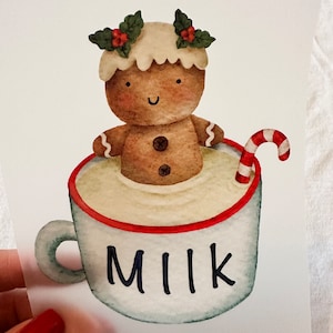 Peut inclure: Un personnage en pain d'épices portant une couronne de houx est assis dans une tasse blanche remplie de lait et surmontée d'une canne en sucre. La tasse porte l'inscription "MILK" sur le côté.